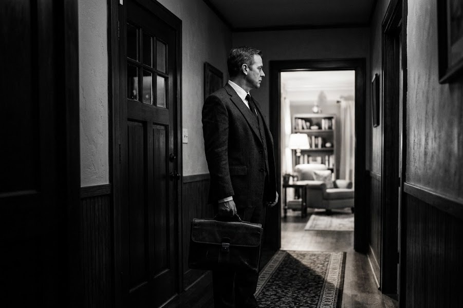 Executive pausing in a dim hallway at night, briefcase in hand, before a lit doorway — the private cost of the chair