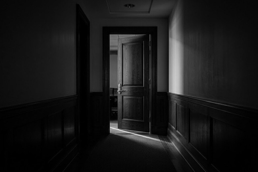 Heavy boardroom door standing ajar at the end of a dark executive corridor, black and white