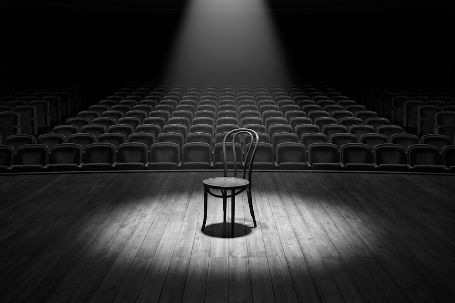 A single chair on a bare stage facing rows of empty auditorium seats, black and white